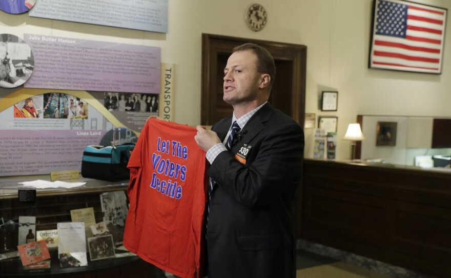 Tim Eyman, a career anti-tax initiative promoter, holds up a t-shirt that reads "Let the Voters Decide," as he talks to reporters, Monday, Nov. 25, 2019, at the Capitol in Olympia, Wash.