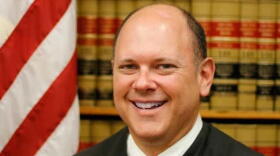 Former state Supreme Court Justice Matthew Rosenbaum in his black judge's robe and tie in front of the American flag and law books
