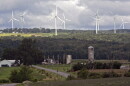 A rural landscape. Farms and trees are visible in the foreground. In the background are 8 large white windmills.