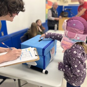 Child wtih pink hat and purple coat puts ballot in a box on a table, with a tabulating person with clipboard attending