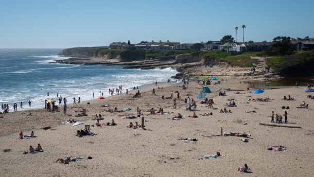 An overview of Natural Bridges State Beach from the upper parking lot shows people lounging on the beach and in the water.