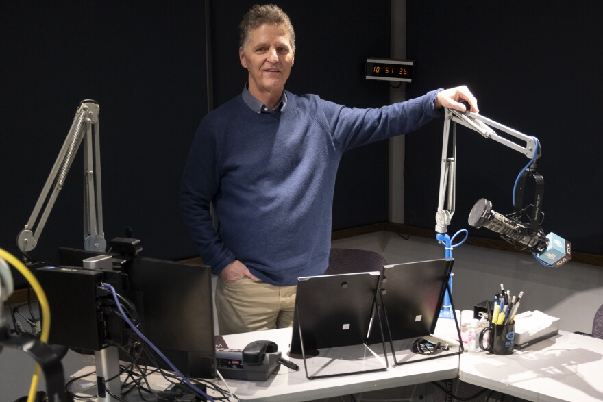 A man in a blue sweater and khaki pants stands behind a desk, resting his arm casually on a studio microphone. 