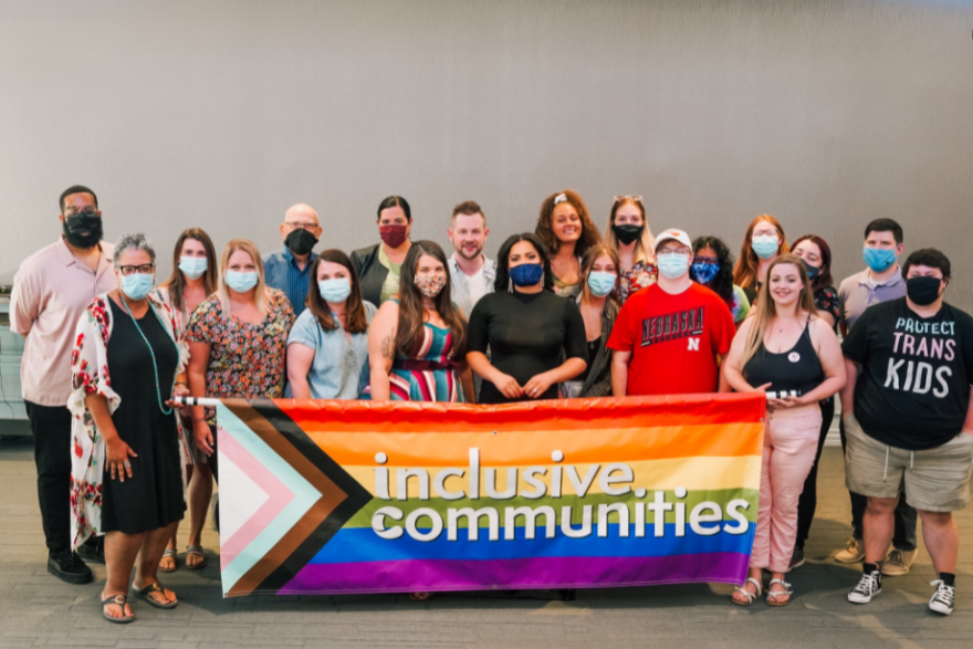 20 people stand in two rows in front of a Pride Flag banner reading "inclusive communities" in white, lowercase text.