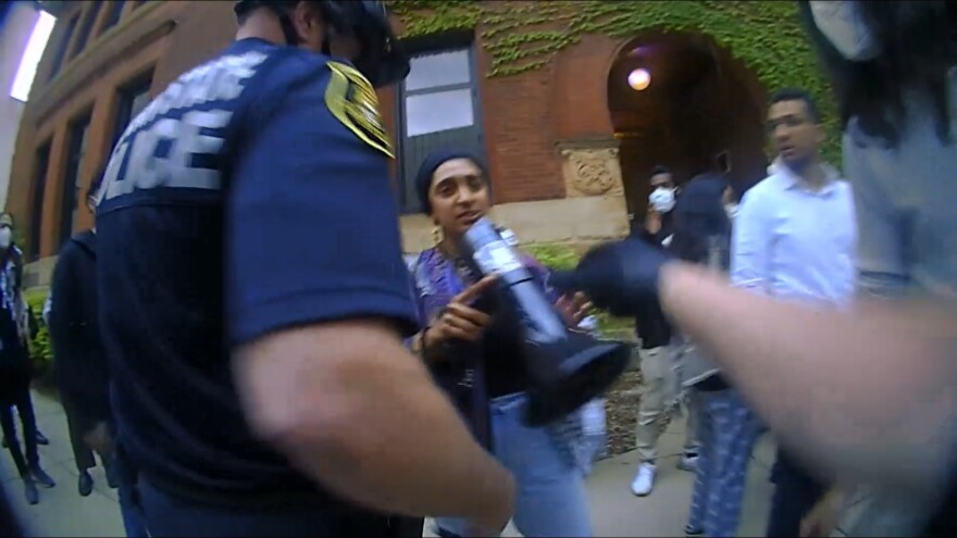 A screen capture from a police officer’s body camera footage shows a police officer reaching out to take a megaphone from a young woman in front of him. They are standing on a sidewalk with a brick building in the background.