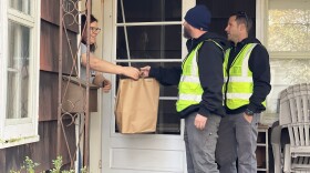 A woman reaches for a brown paper bag from her front door as two public works staff standing on her porch hand it over to her.