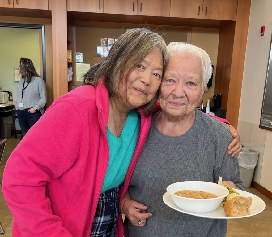 Two women stand close together, one holding a plate with bread and a bowl of soup.