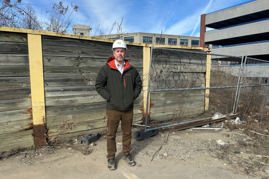 A man wearing a white hard hat stands outside a fenced area