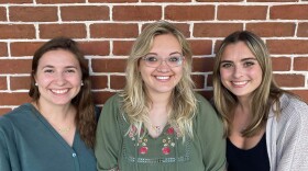 Three young students stand in front of a brick wall and smile at the camera.