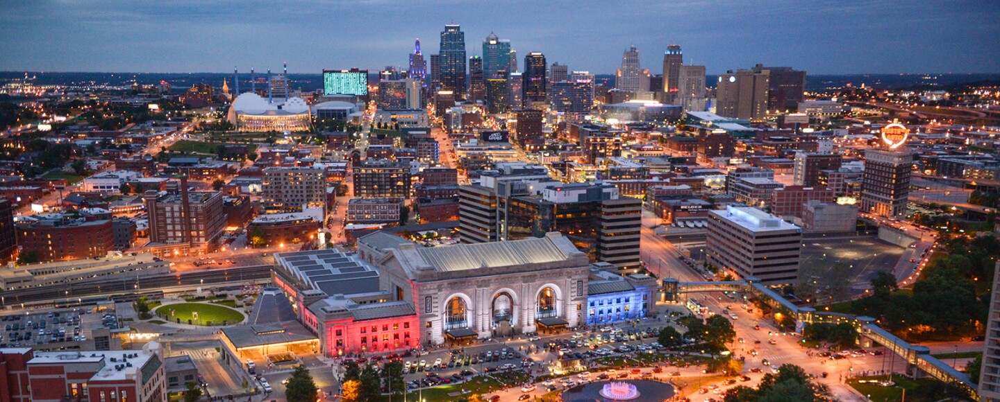 A wide view of the skyline of Kansas City shows several hot air balloons in the foreground and the cityscape extending into the background. It's a late evening hour and streetlights illuminate the roadways with an orange glow.