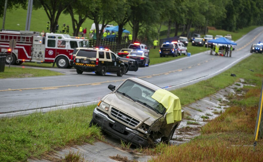 A brown Ford pickup truck with damage to its right side is parked on the ride of a road. On the other side of the road are police cars and fire trucks. In the distance are emergency personnel gathered around an overturned white bus. 