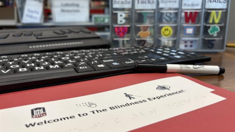 A red folder sits on a desk, surrounded by a marker, a large-print computer keyboard, and a storage bin that visually-impaired people can use for hardware. One of the rows is labeled "nails" and features a textured letter "n" and an actual nail glued onto the front so they could distinguish what's placed inside. The red folder reads "Welcome to the Blindness Experience!" and says the same underneath in braille. 