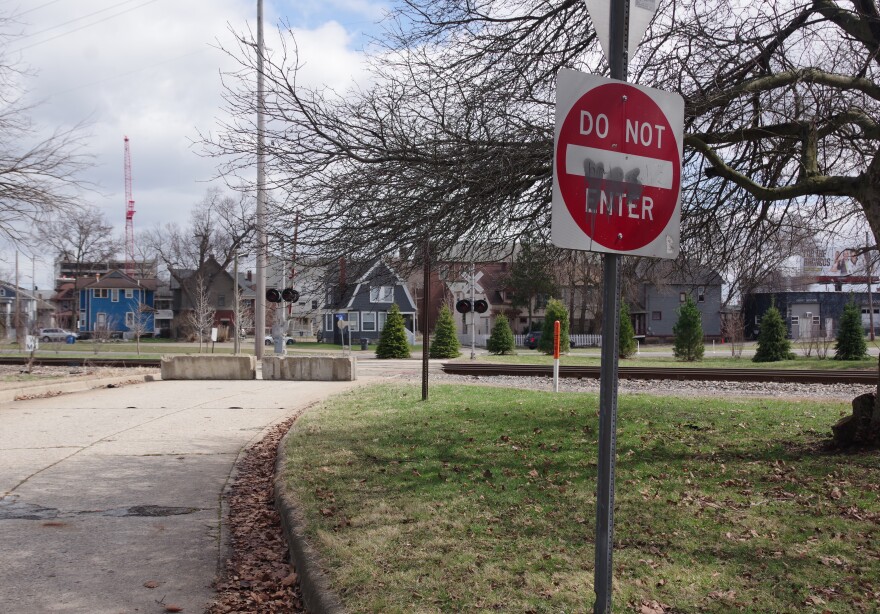 A partially defaced Do Not Enter sign sits at the end of a short gray road, which can be seen crossing over rail tracks in the distance. However, concrete blocks stop any through traffic from entering the road, with the rest of it beyond the blocks covered by grass and two young pine trees.