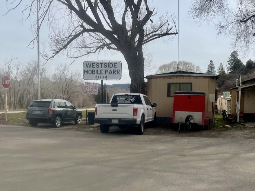 The entry of Westside mobile home park. There is a road to the left of the composition with two mobile homes to the right in full or partial view. Two cars are parked near one of the homes.