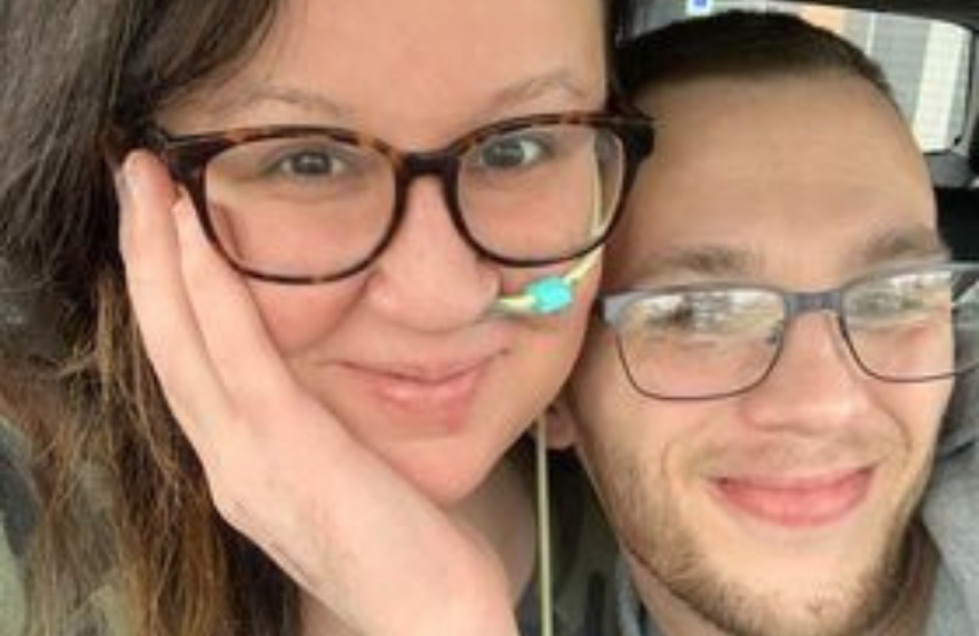 A couple smiling at the camera. The woman has a feeding tube.