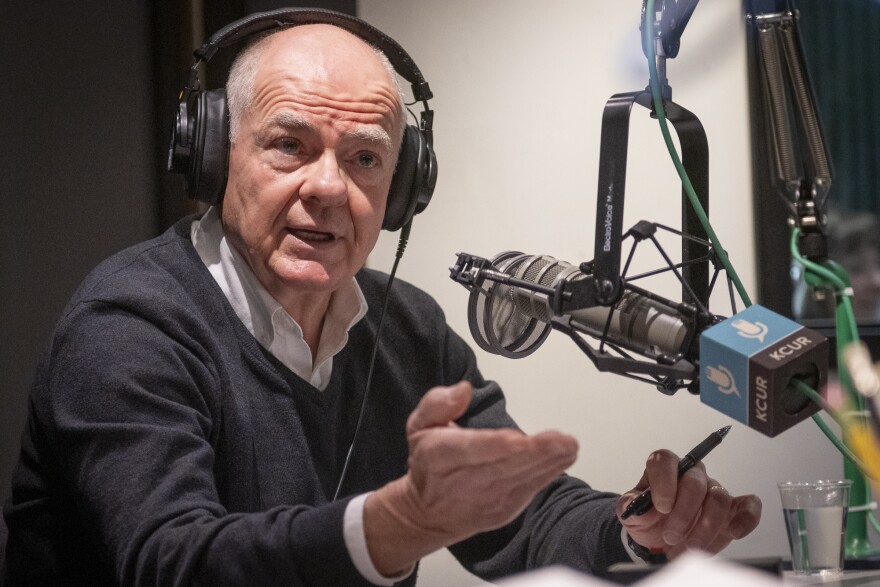 A man inside a radio studio sits behind a microphone talking and gesturing with his right hand.