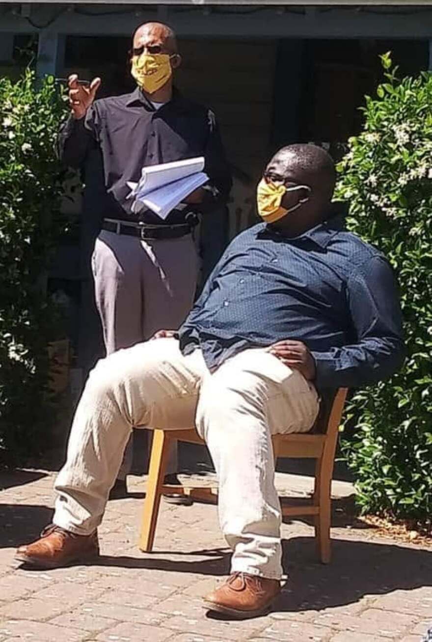 One man talking, another man seated. 