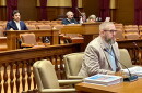 A man with a beard and glasses sits at a table in a courtroom, with other people seated in the background.
