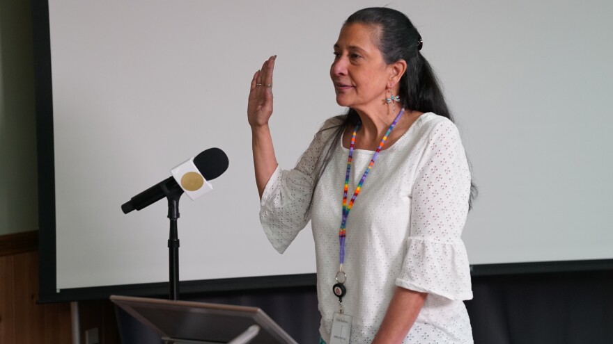 A woman in a white blouse stands at a podium, right in front of a large white projector screen. She raises her right hand, palm and fingers stretching out flat as she looks to the left side of the photo. A mic can be seen held by a mic stand next to her raised hand, with a white pyramid style mic flag with a gold circle in the middle of one edge of the flag. The woman is also wearing a long lanyard made of rainbow colored beads. 