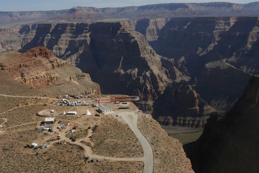 This is an aerial view of part of the Grand Canyon which features astonishing large mesas and cliffs lining a large canyonland area. This view also shows us the Skywalk, a large u-shaped walkway that goes out over the canyon and allows people to have a rare and unique view of the canyon and river below. There are some tipis and other structures visible 