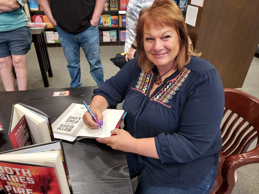 Bobbie Scopa smiling at the camera while she signs a copy of her book "Both Sides of the Fire Line" at a bookstore.