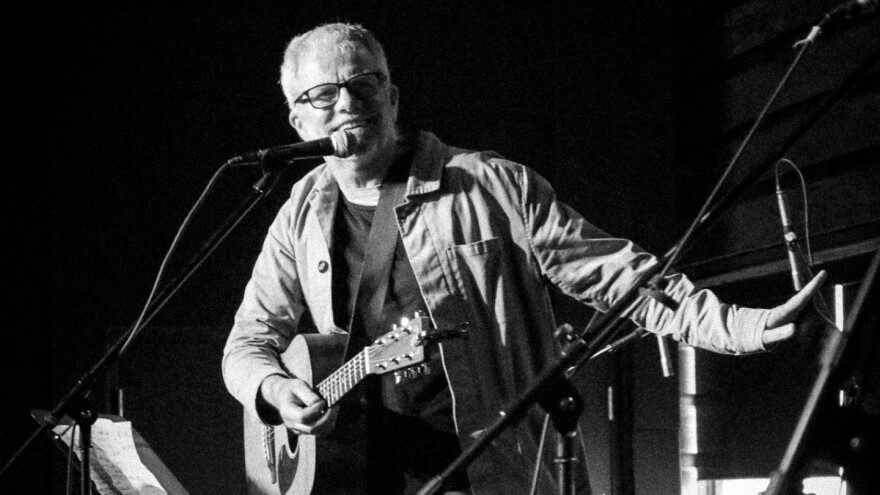 A man wearing glasses and holding an acoustic guitar smiles on stage in a small concert venue.