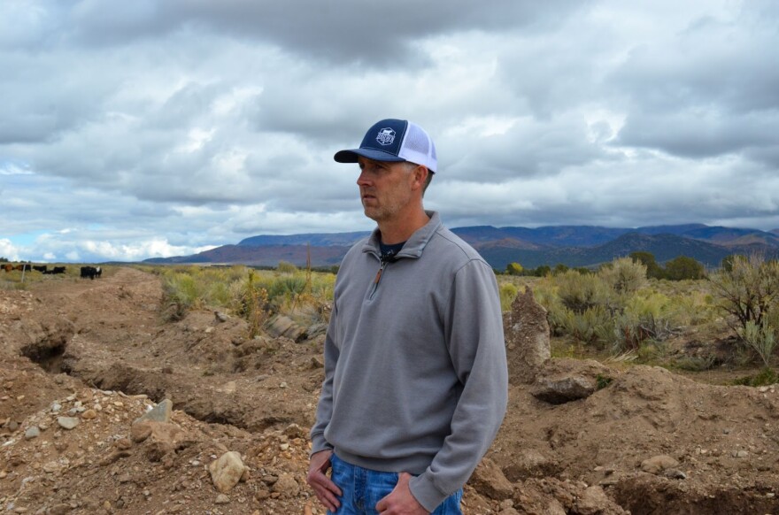 A man in a gray sweater and baseball cap stands in front of an irrigation ditch. Mountains and shrub are in the background.