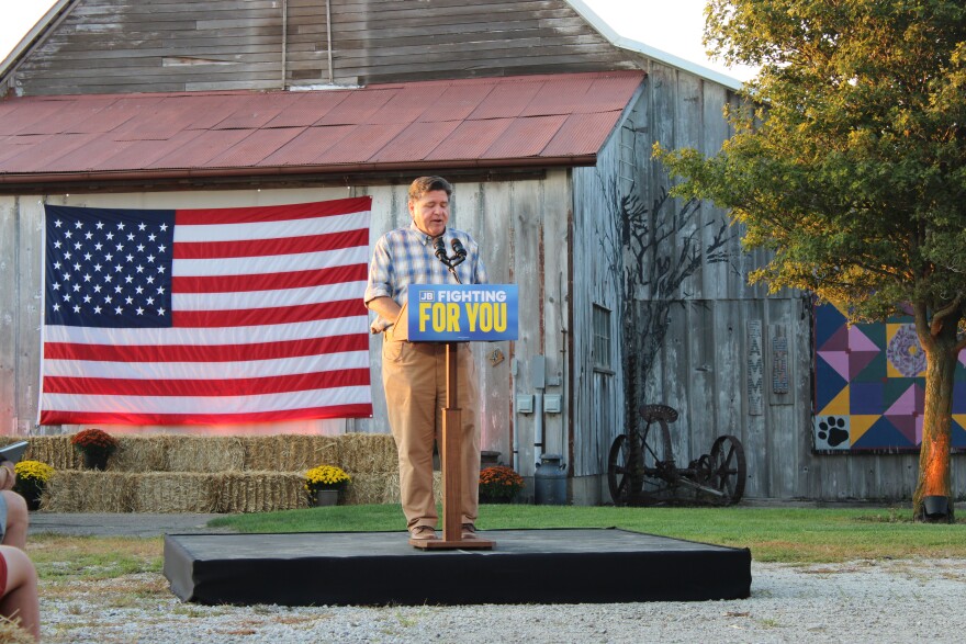 Gov. JB Pritzker speaks at a podium labeled “Fighting for You” in front of a barn with a large American flag, hay bales, and a tree nearby.