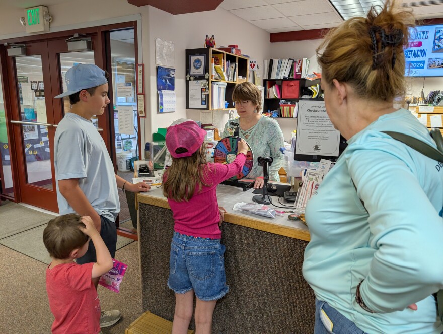 Delta Community Library Director Tiki Levinson, behind counter, helps another family get rewards for reading. Ten-year-old Reilly Testa takes a turn at the prize wheel for reading several books as part of the Delta Community's Library's Summer Reading Program. Her brothers, Mikey, 3, and Peter, 12, wait for their turn as their grandma, Kat Connor, looks on.