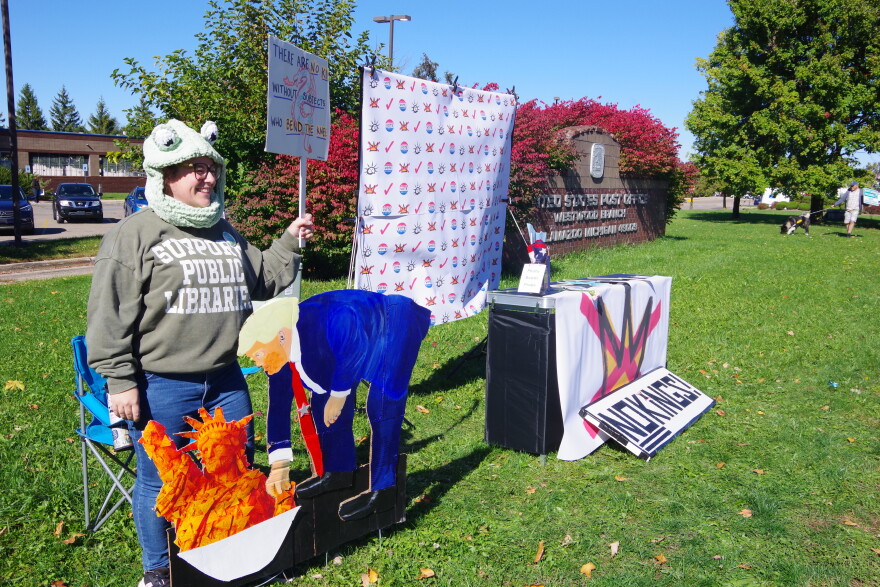A woman in a knitted frog hat and holding a sign stands behind a cut out poster of President Trump dipping into a Statue of Liberty made out of Doritos