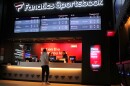 A man places a bet at the counter at a retail sportsbook as three screens overhead display game stats. 