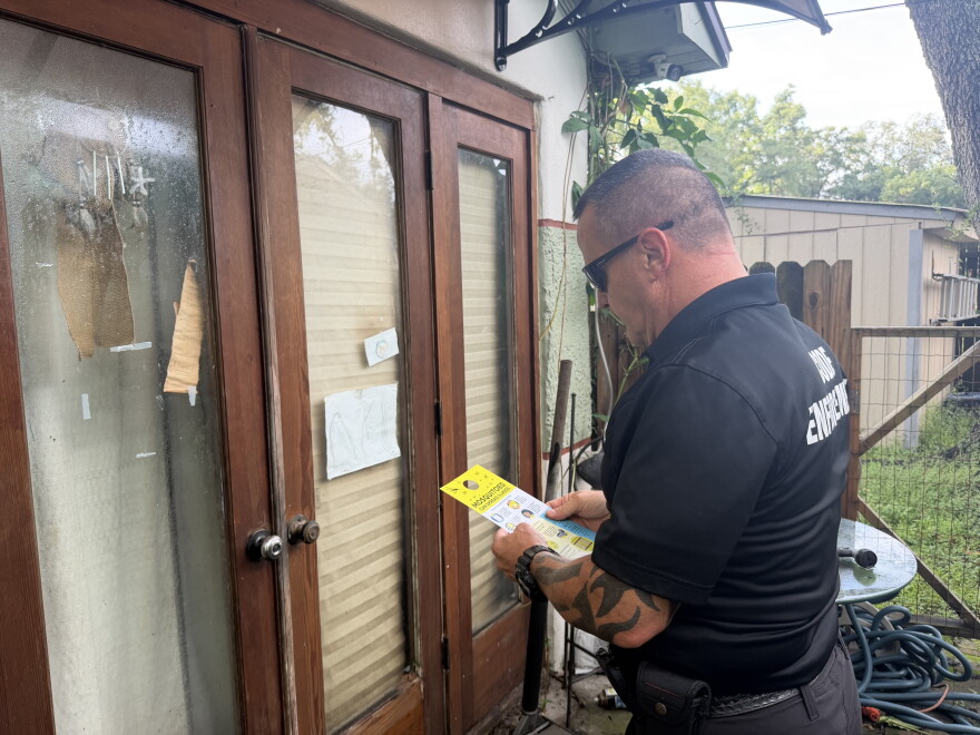 A man wearing a black polo that reads "code enforcement" on the back, stands looking down at a yellow flyer. He is in front of a brown door with tan shutters and a broken window. There is another house, grass, and a fence in the background.