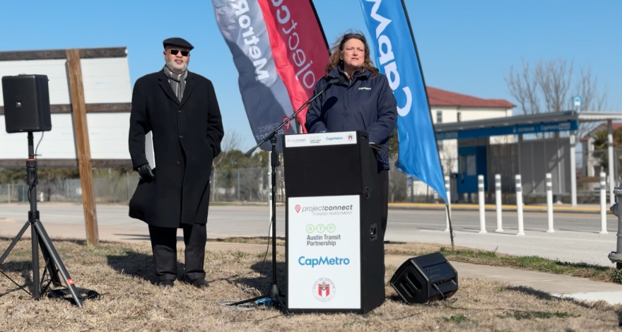 A woman in a jacket is standing at a podium that says, "Project Connect," "Austin Transit Partnership," and "CapMetro." Behind her stands a man dressed in black wearing sunglasses, a scarf and hat. Public address speakers are standing. Behind them are vertical CapMetro flags fluttering in the wind.