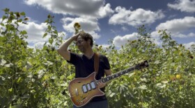 A man wearing a short-sleeve shirt and holding an electric guitar runs a hand through his hair while smiling in front of tall, wild plants and a blue sky with white clouds.