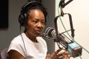 A woman inside a radio studio sits behind a microphone talking. She is gesturing with both hands.