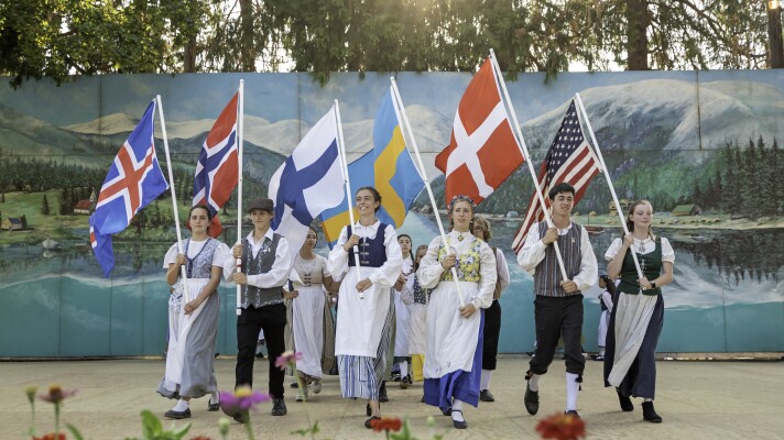 People on stage dressed in 1800s Scandinavian clothing, holding flags from each Scandinavian country. 