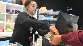 A woman, left, wearing a black jacket hands cash to a person whose arm we see at left. They are making the exchange over a counter. Behind the woman are various cannabis products on a shelf.