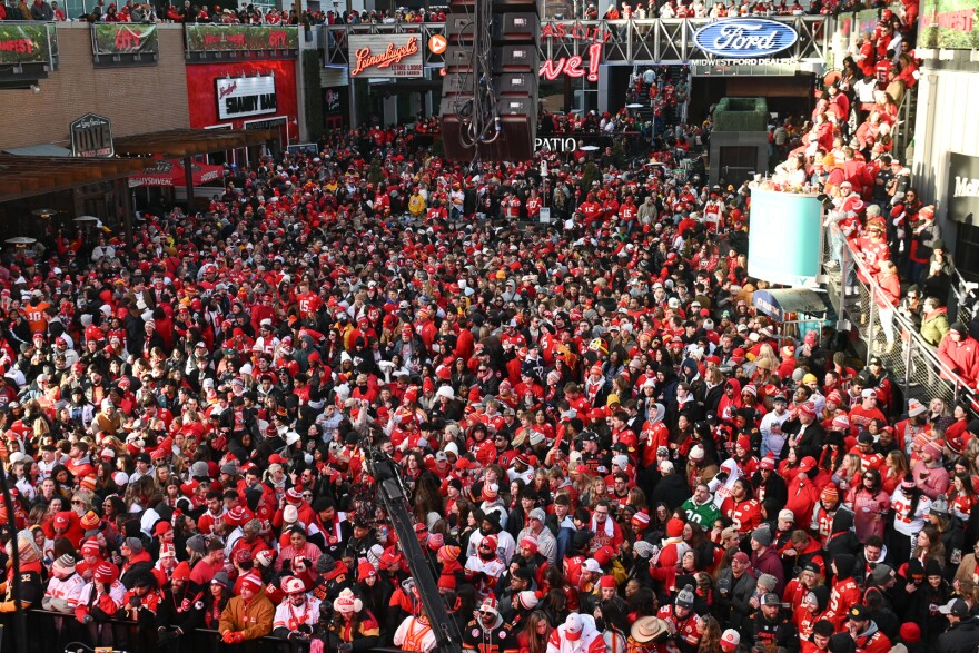 A crowd of thousands pack together inside a venue. There are many lighted signs for restaurants around and above the crowd.