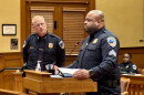 A police officer stands at a podium speaking, while another officer stands behind him and a third officer sits at a desk in the background.