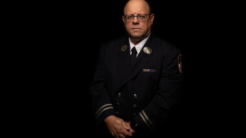 A middle aged man in a firefighter's dress uniform stands in front of a black background with a somber expression.