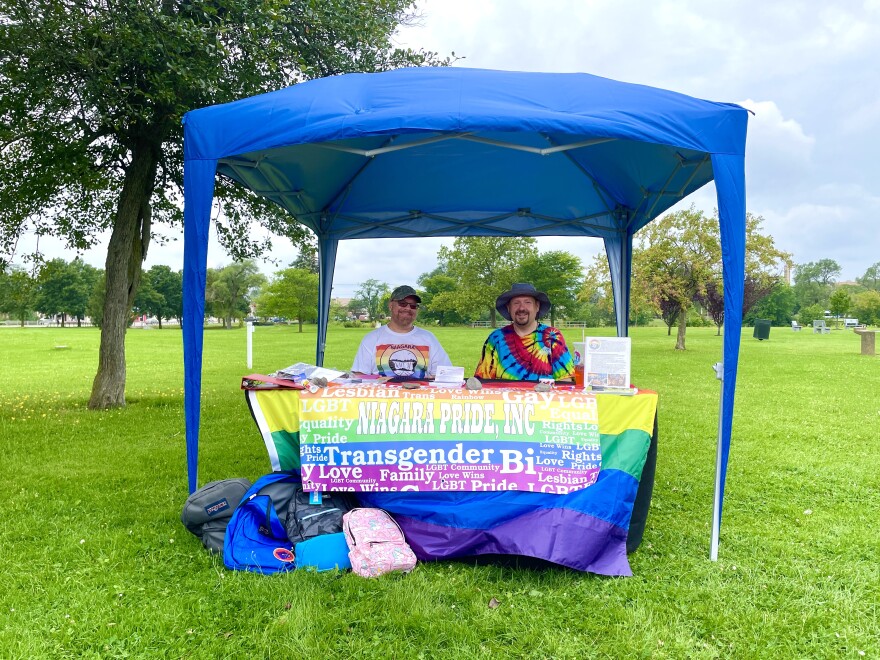 Ronald Piaseczny and Mark Smith sit at Niagara Pride's donation table under a blue gazebo tent in Hyde Park. The table has backpacks sitting in front of it that were donated on Friday and has two rainbow flags hanging on the front. 
