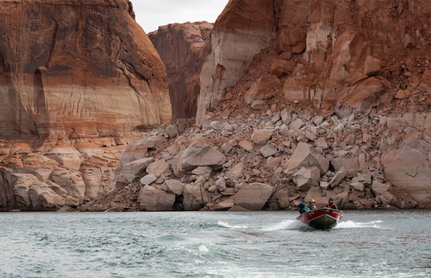 A small boat skims across the blue water of a lake. Behind the boat are large cliffs with white markings up to the middle of the cliff. Above the white markings, the cliffs are orange.