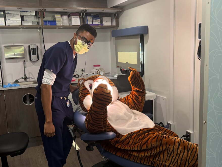 A dental professional wearing scrubs, a mask and protective glasses examines a person in a tiger mascot costume seated in a dental chair.