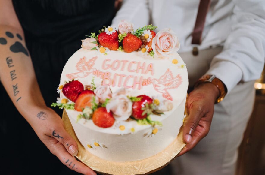 A single-layer, white wedding cake is seen from above. It's decorated with strawberries, fresh roses and the words, "gotcha bitch."