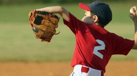 Young baseball player in a red jersey winds up to pitch, wearing a glove and cap, on a dirt infield with green outfield grass in the background.