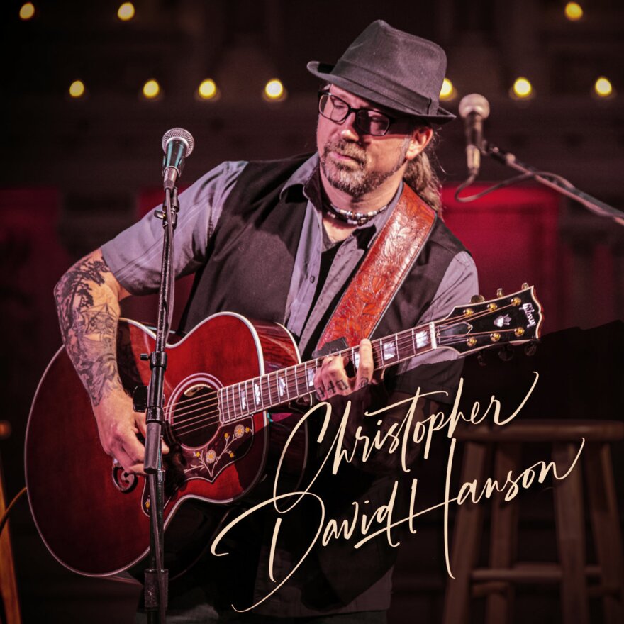 A man holding a red acoustic guitar standing behind a microphone. "Christopher David Hanson" is written over the photo.