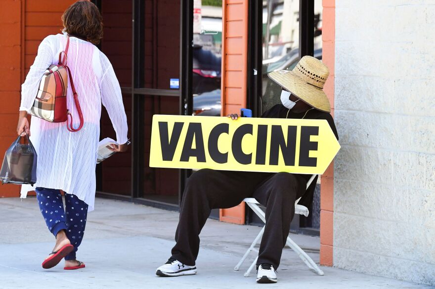 A person sitting down on the sidewalk holds a yellow sign reading "VACCINE" as a woman passes by.