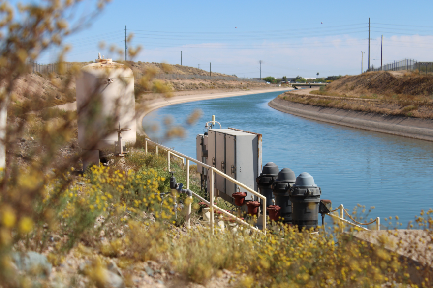 A canal flowing in a dry area. Beside the canal is a tank and some electrical infrastructure.