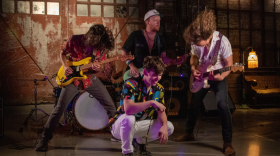 A singer in a colorful shirt squats on the ground while laughing as three guitarists stand behind him and play.