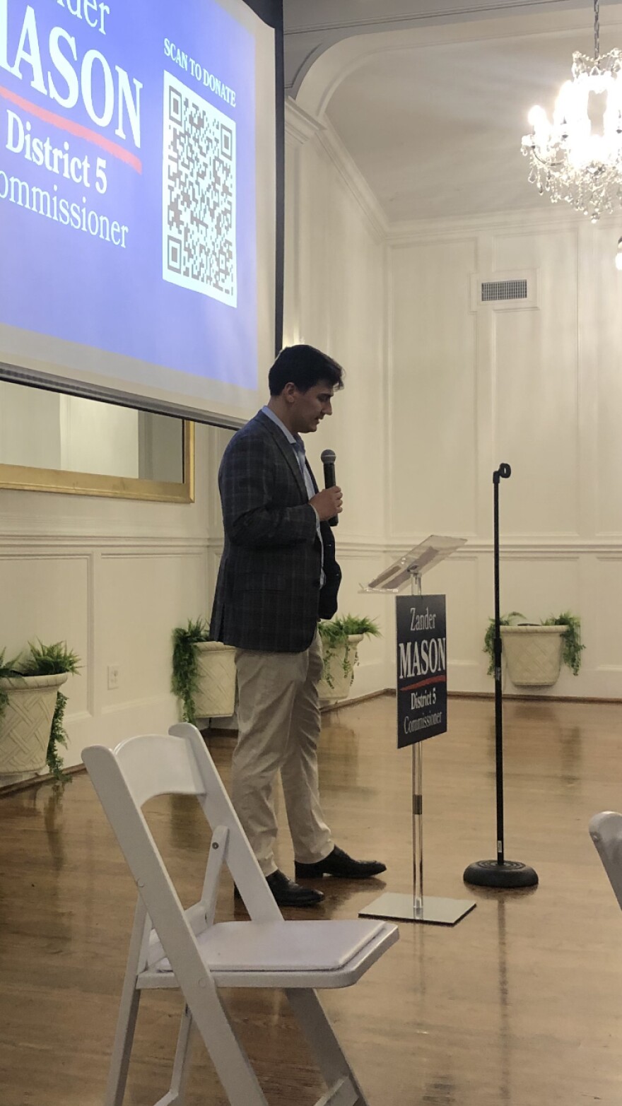A man stands at a podium with a microphone, speaking at an indoor campaign event.