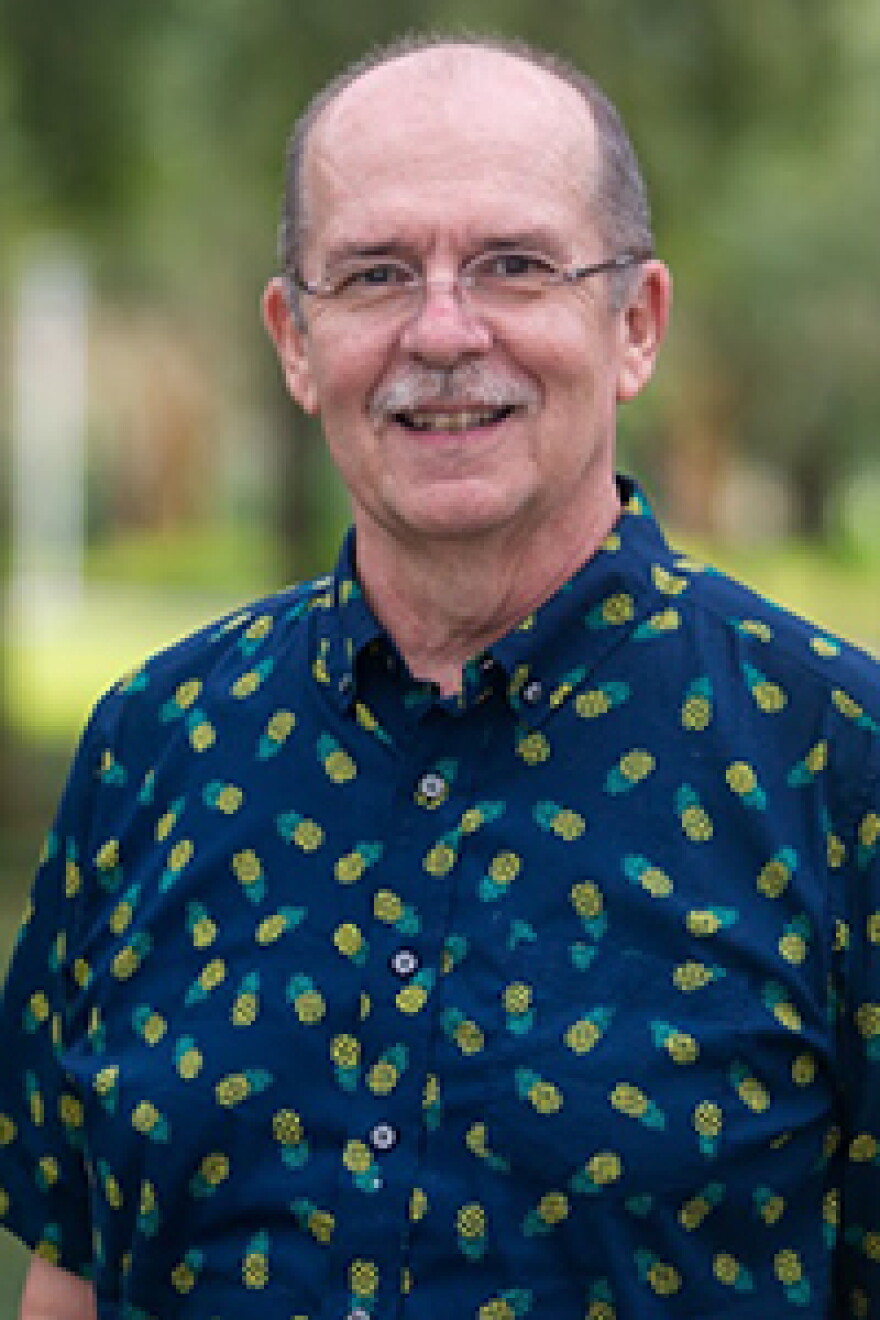 Bald man with a gray mustache and glasses wearing a blue short-sleeve button-down shirt and smiling into the camera